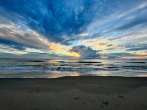 A serene beach at sunrise with soft waves rolling onto the sand, golden light illuminating dramatic clouds, and a pier extending into the water on the right.
