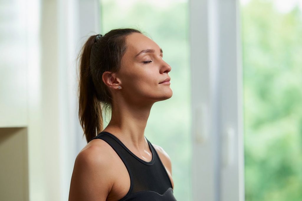 Woman practicing slow diaphragmatic breathwork with eyes closed beside a bright window with green trees outside
