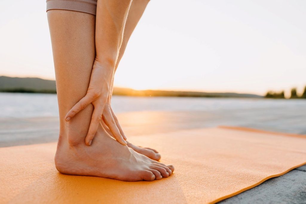 Bare feet resting on an orange yoga mat beside a lake at sunset, one hand touching the ankle, illustrating a feet-to-floor somatic grounding techniques.