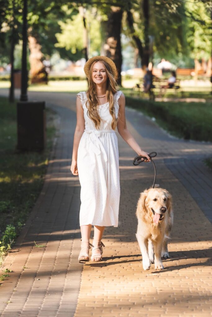 Woman walking a golden retriever along a sunny park path in a relaxed outdoor setting.
