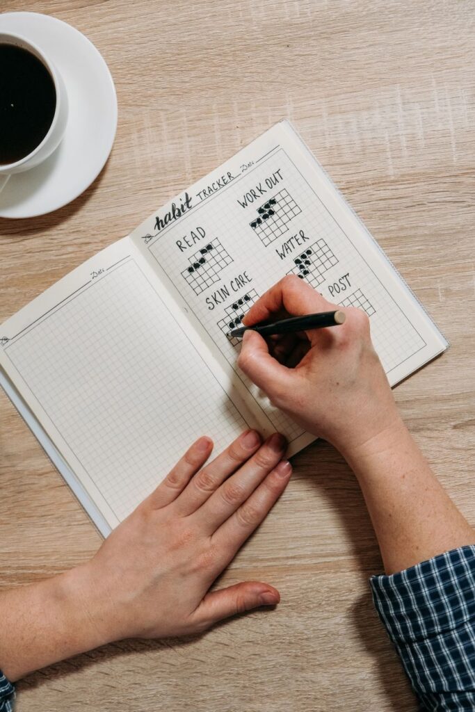 Hand marking a habit tracker notebook beside a cup of coffee on a wooden table.