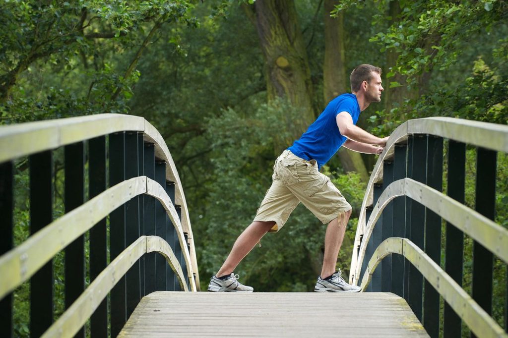 Man doing a calf stretch on an outdoor bridge surrounded by trees, part of a morning mobility routine.