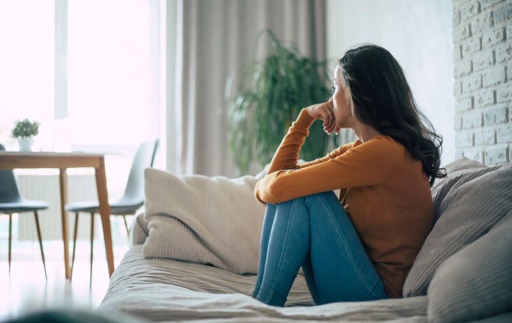 Woman sitting curled up on a couch in a quiet living room, looking toward the window, capturing a moment of nervous system dysregulation and withdrawal.
