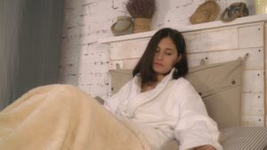 Woman sitting in bed wearing a white robe, looking tired and fatigued, wrapped in a blanket in a calm bedroom setting.