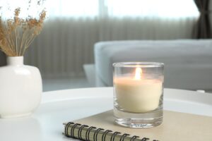 Lit candle on a notebook beside a ceramic vase with dried stems in a softly lit, calming bedroom.