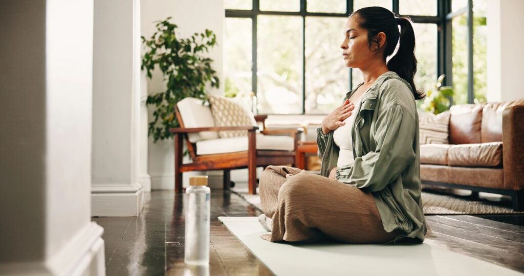 Woman sitting cross-legged on a yoga mat in a bright living room, eyes closed and hand on chest, practicing mindful breathing or meditation.