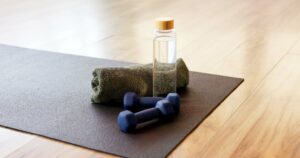 Minimalist workout still-life with a yoga mat, small dumbbells, a rolled towel, and a clear water bottle on a wooden floor, representing gentle movement and active aging.