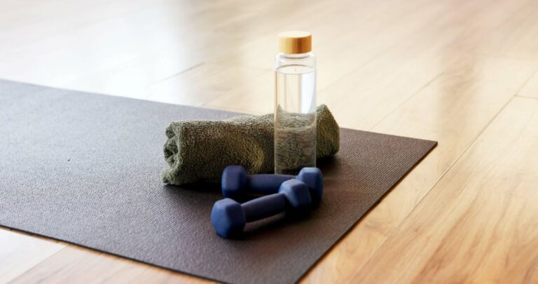 Minimalist workout still-life with a yoga mat, small dumbbells, a rolled towel, and a clear water bottle on a wooden floor, representing gentle movement and active aging.
