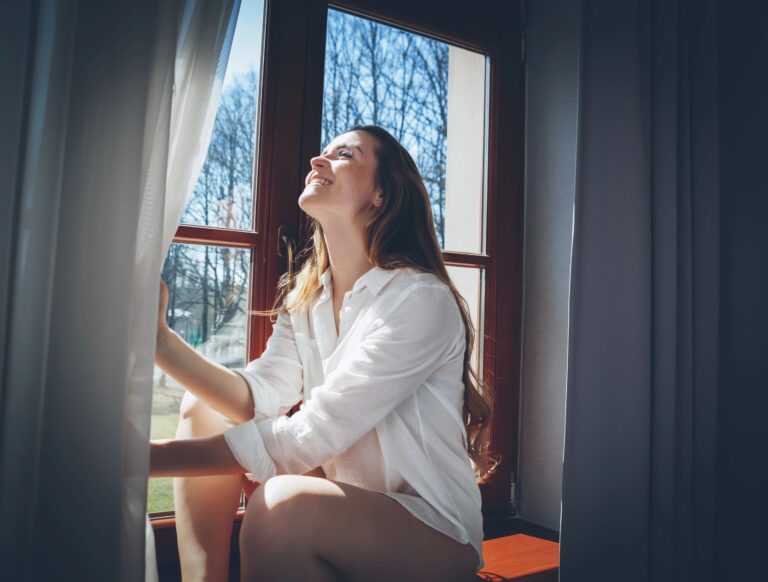 Person sitting on a windowsill opening a curtain as natural light streams in on a clear morning.