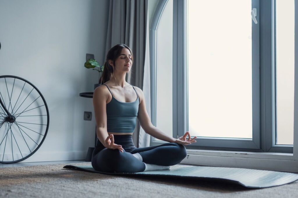Person meditating cross‑legged on a yoga mat near a bright window