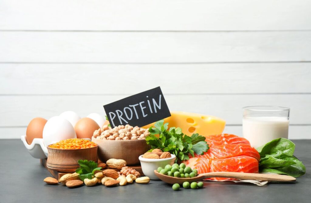 Assorted protein‑rich foods arranged on a dark surface, including salmon, eggs, legumes, nuts, cheese, and milk with a small sign labeled protein.