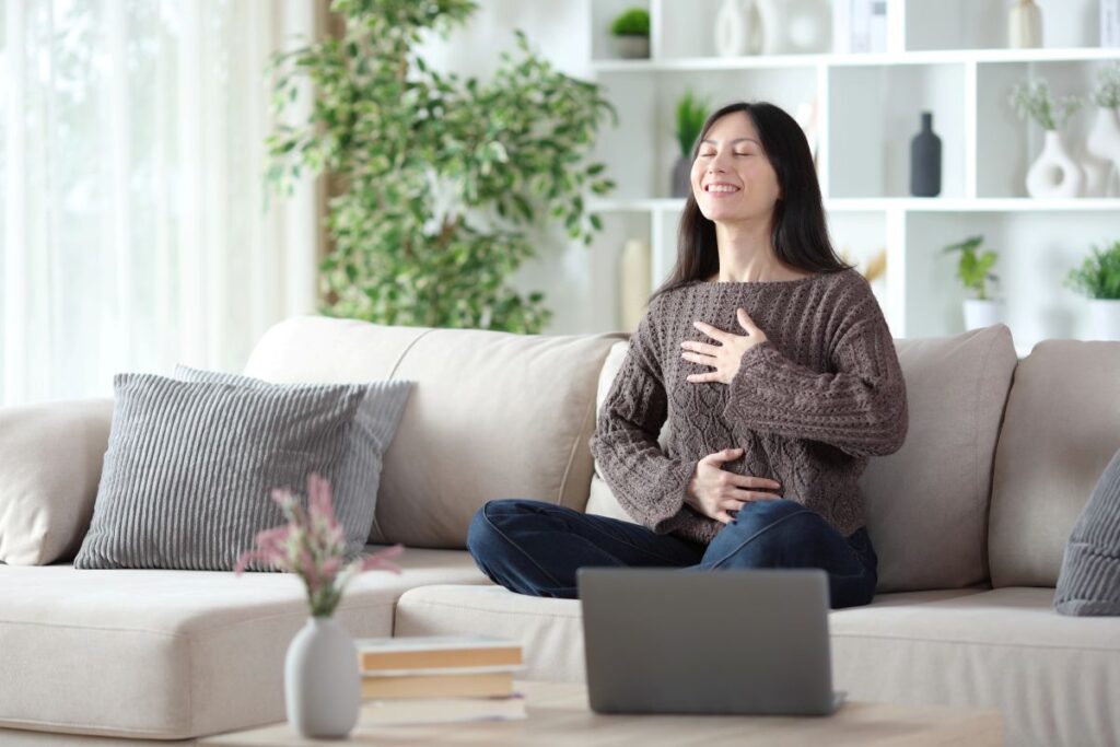 Woman sitting on a sofa practicing a calm somatic check‑in with one hand on her chest and one on her abdomen in a cozy, sunlit living room with plants and soft neutral decor.