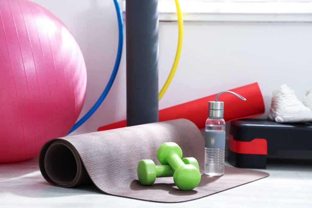 Home workout equipment arranged in natural light, including a pink exercise ball, yoga mat, dumbbells, water bottle, athletic shoes, and step platform.