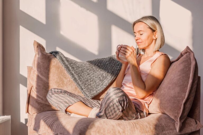 Woman with eyes closed holding a warm cup in a sunlit chair, a quiet recovery pauses for cortisol and cognitive reserve