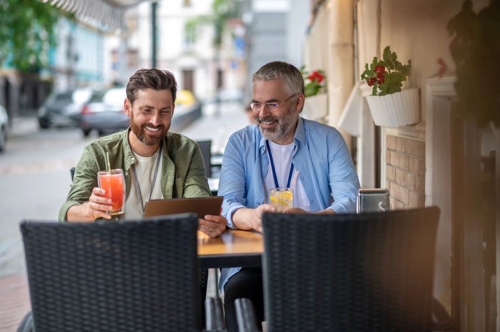 Two smiling men sharing drinks and a tablet at an outdoor café, an everyday form of social connection